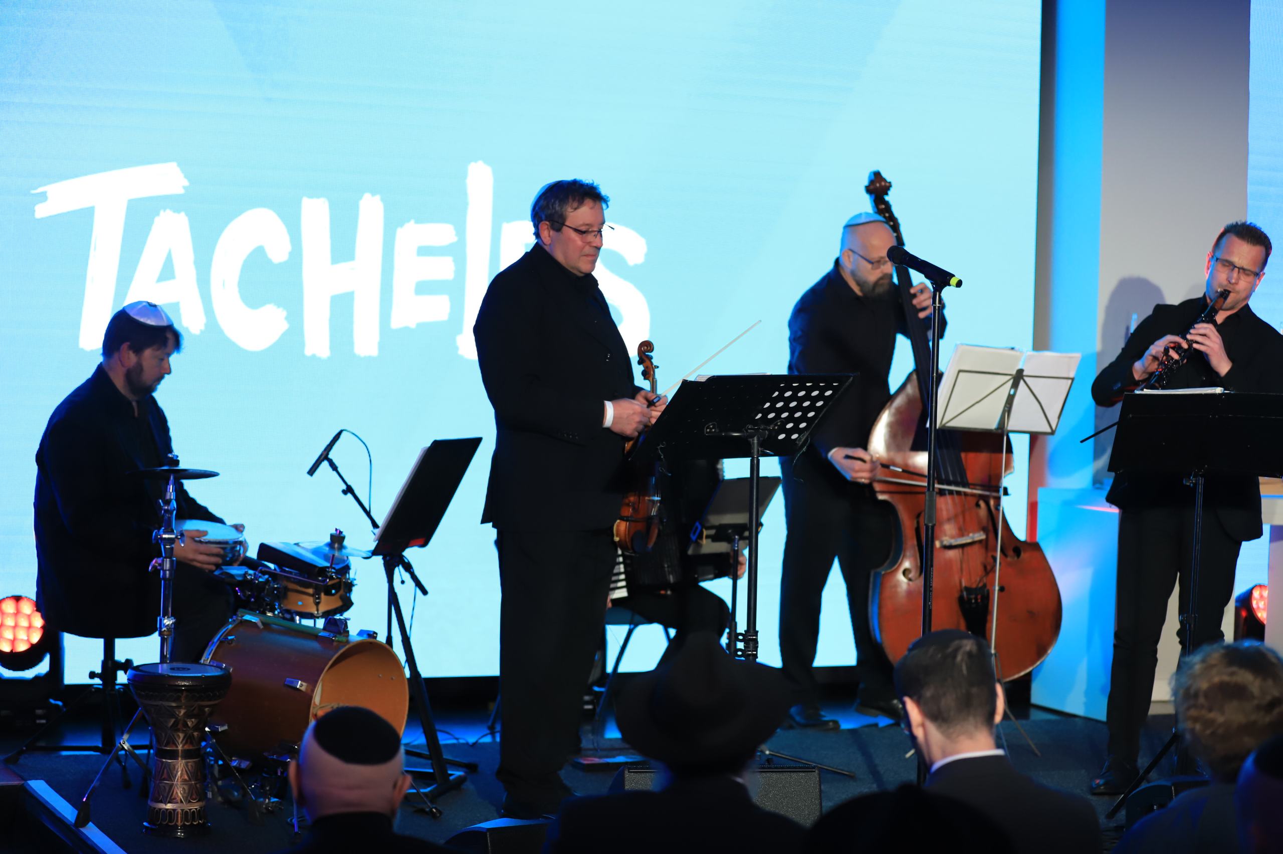 Vier Männer in schwarzen Anzügen spielen auf einer Bühne, die Instrumente sind Schlagzeug, Geige, Kontrabass, Klarinette