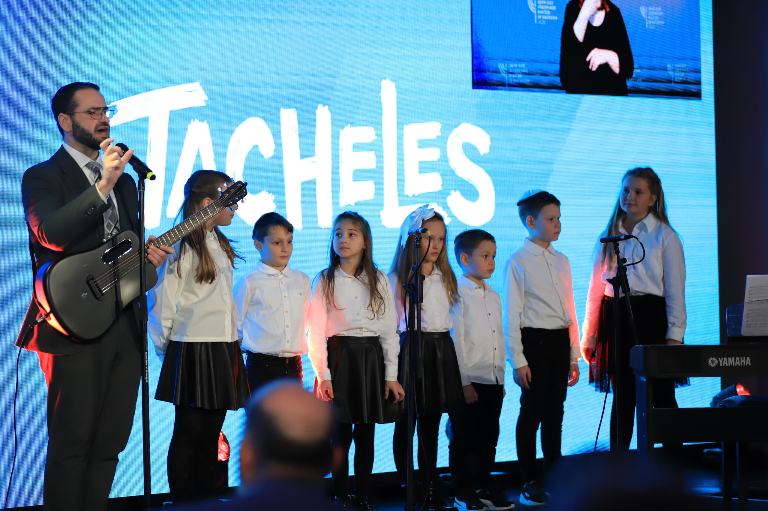 Sieben Kinder in weißen Hemden stehen auf einer Bühne, neben ihnen ein Mann mit Gitarre