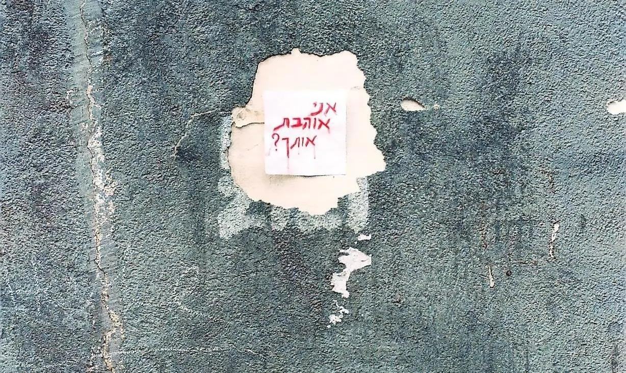 Ein Post-it Zettel an einer Wand. Darauf "Liebe ich dich?" in Hebräisch.
