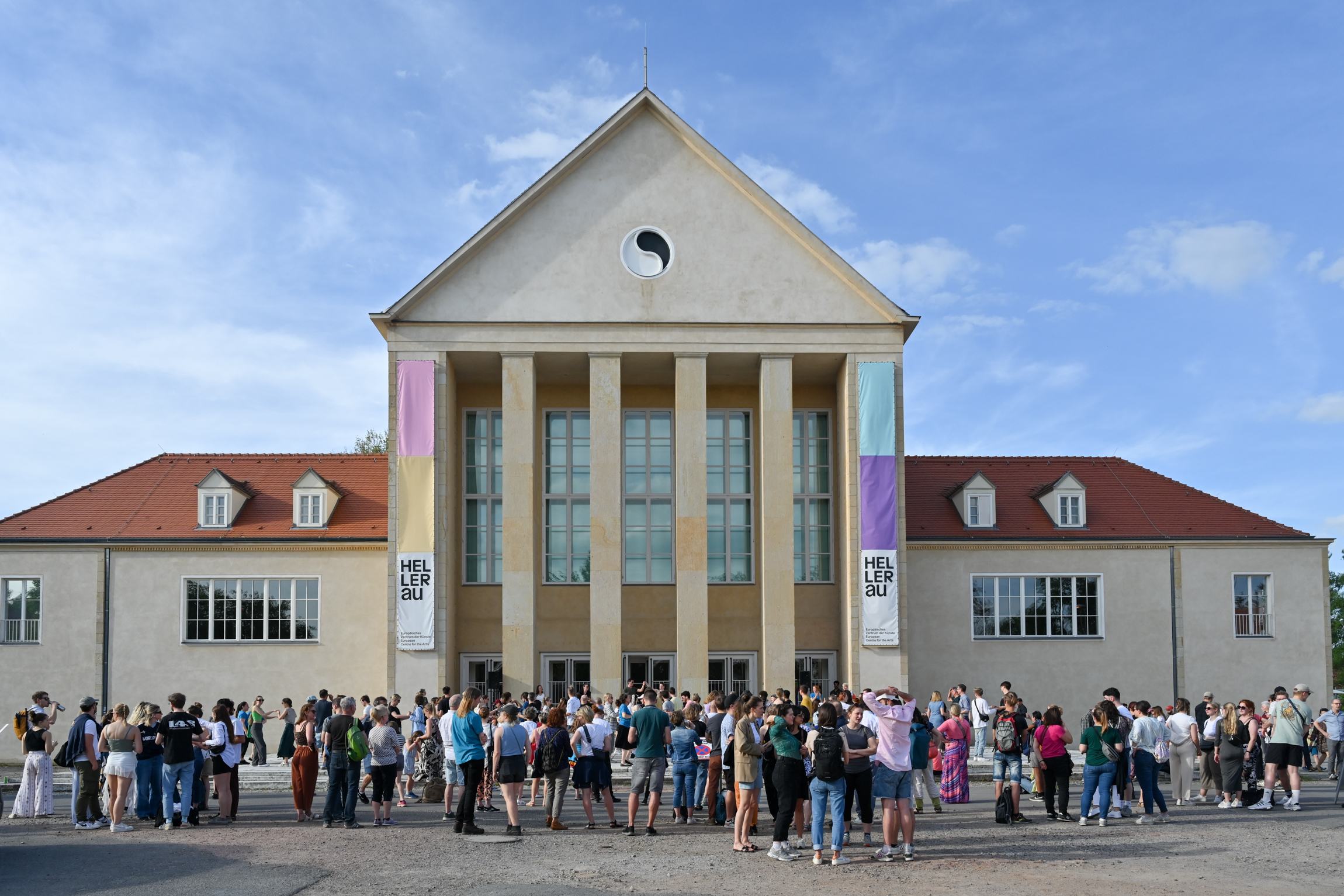 Große Menschenmenge vor einem großen Gebäude mit Säulen und roten Dächern unter blauem Himmel.