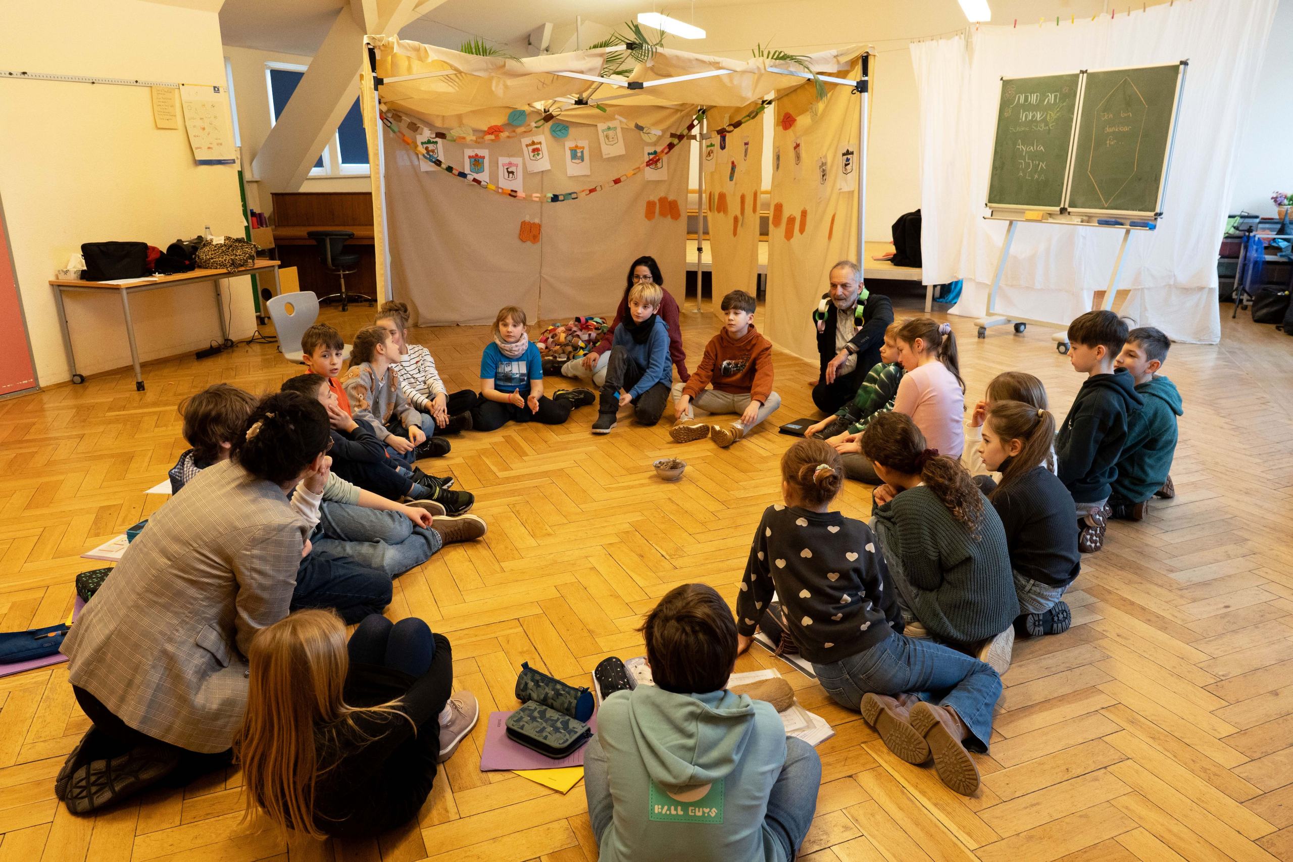 Kinder sitzen in einem Kreis vor der selbstgebauten Laubhütte (Sukka)