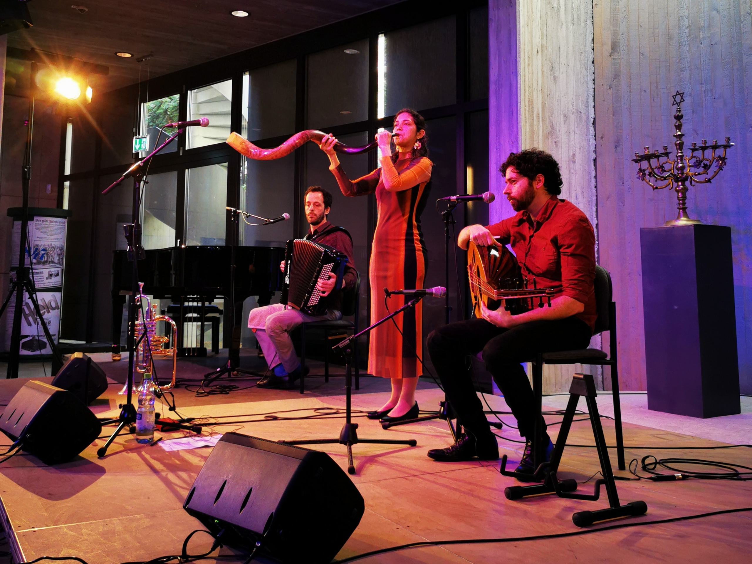 Ein Konzert der Band Folkadou in Leipzig. Man sieht zwei Männer, sitzend, die Akkordeon und Oud spielen, und in der Mitte die SÄngerin, die gerade das Schofar, ein Widderhorn, bläst.