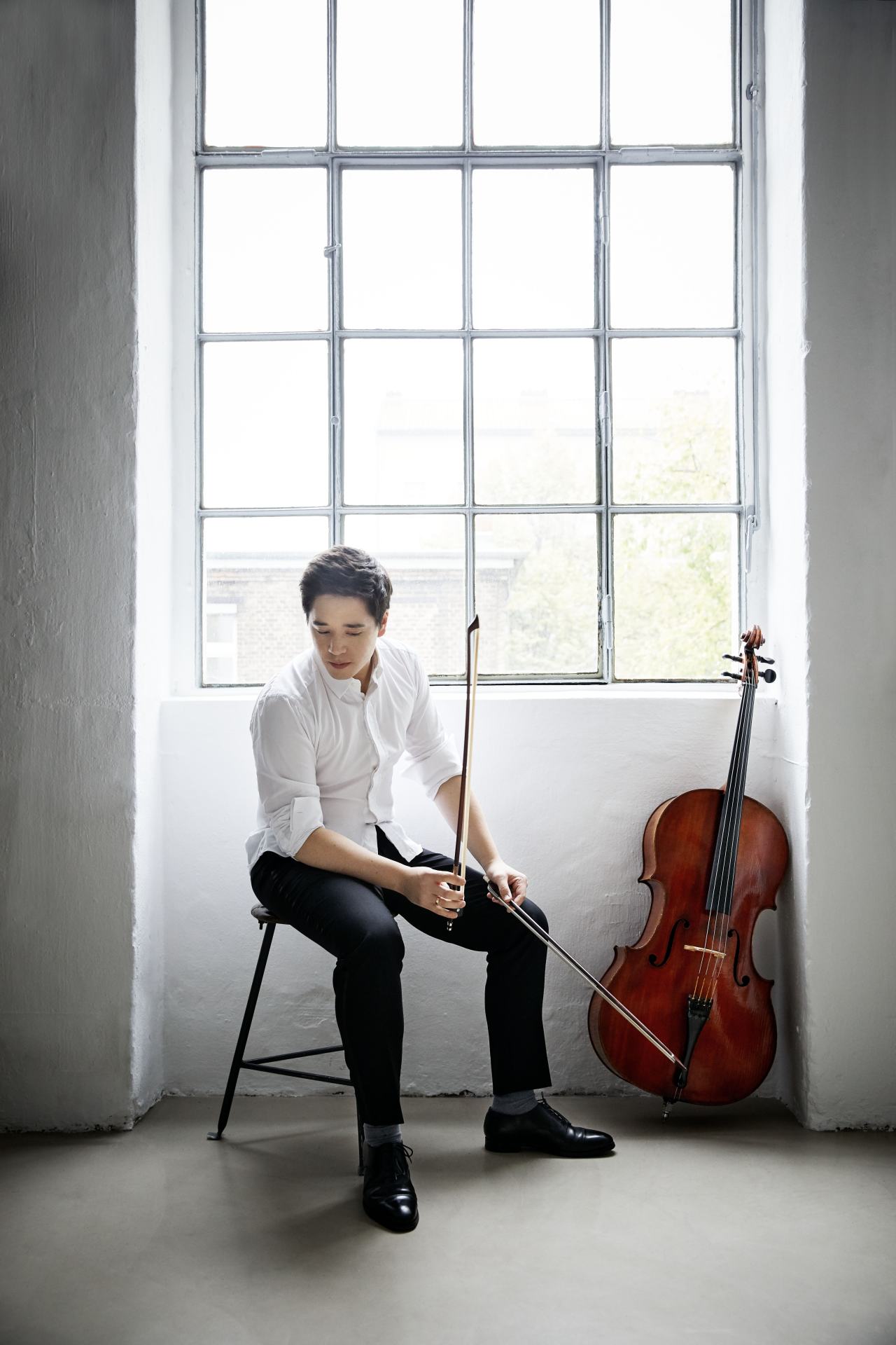 Ein Cellist sitzt mit seinen zwei Cello-Bögen auf einem Stuhl vor einem Fenster und einer weißen Wand. Er trägt eine schwarze Hose, schwarze Lackschuhe und ein weißes Hemd. Sein Cello lehnt nebem ihm an der Wand.