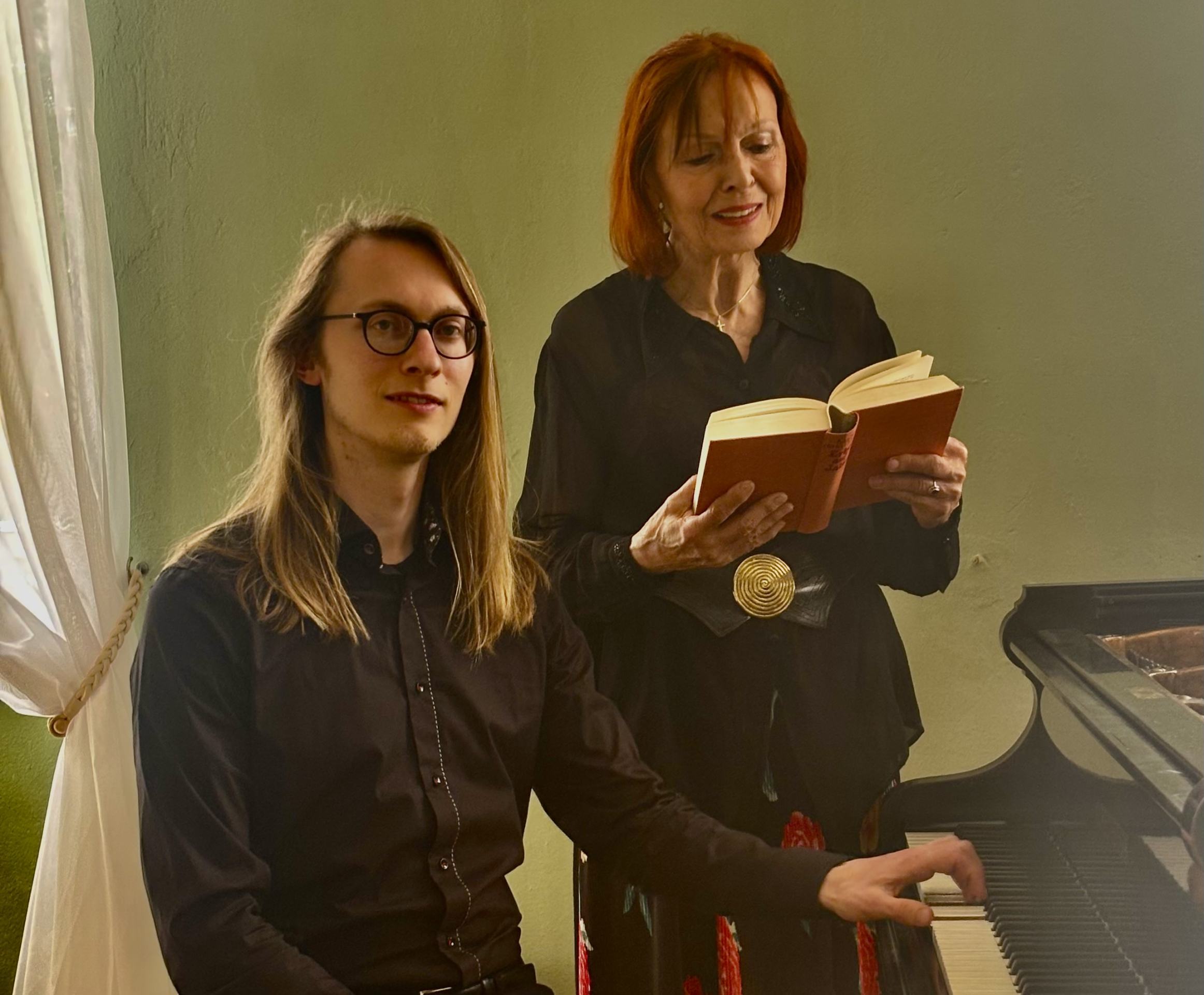 ein junger Mann mit langen Haaren und Brille sitzt am Klavier. Neben ihm steht eine Dame mit halblangen roten Haaren, die ein Buch in der Hand hält