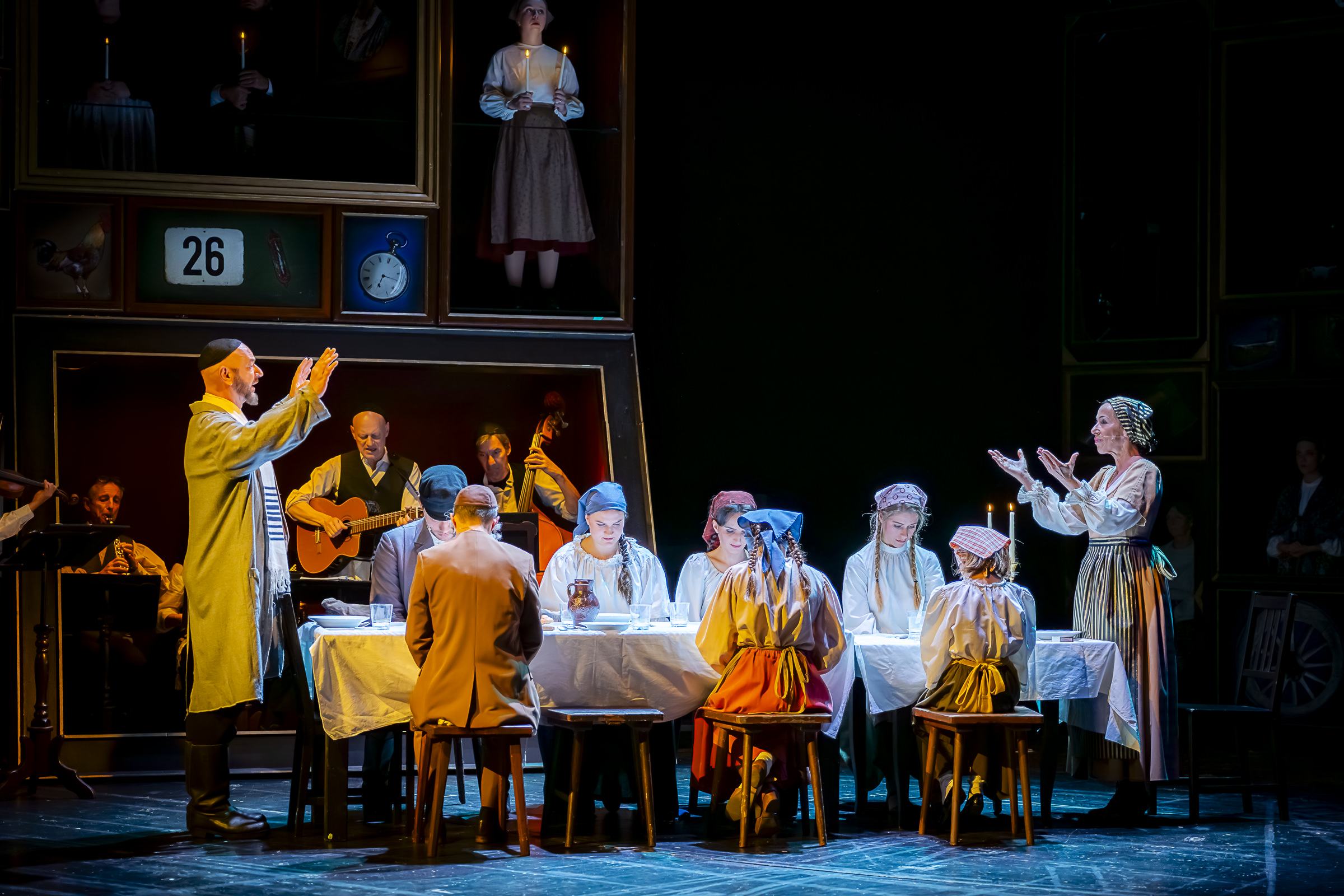 Eine theatralische Bühnenszene zeigt Schauspieler, die um einen langen Tisch versammelt sind zum Essen. Der Mann und die Frau an den Stirnseiten heben die Hände zum jüdischen Tischgebet. Die anderen sitzen mit gesenkten Köpfen.  Gerahmte Bilder und Menschen in Kostümen säumen den schwach beleuchteten Hintergrund